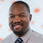 A man with short hair and a trimmed goatee smiles while wearing a light gray dress shirt and a striped tie. The background is white with orange blurred spots.